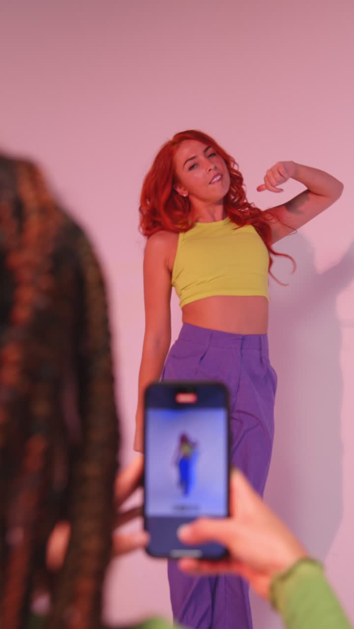 video studio verticale di una donna che scatta una foto di un'amica che balla sul cellulare contro uno sfondo rosa 2