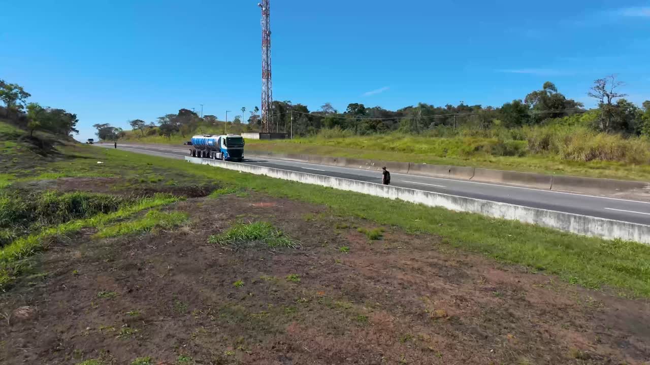 vista panorámica de una carretera rural con un peatón caminando a su lado y una torre de comunicación en el fondo.