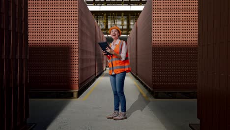 full body side view of asian female engineer with safety helmet looking at the tablet in her hand and looking around while standing with red brick packed in stacks are stored