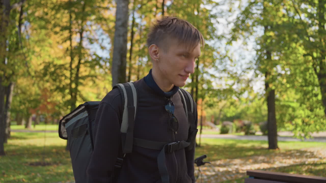 White courier with backpack in park, young white male wearing black jacket adjusts straps of insulated delivery bag at wooden bench, autumn trees and dappled sunlight create warm atmosphere, focused