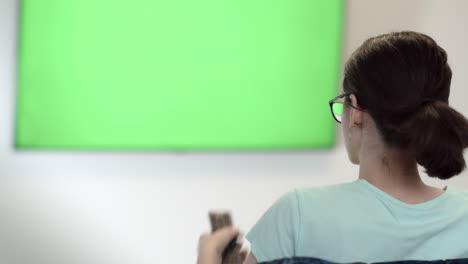 brunette girl sits in font of a geen sreen tv, switches channels, attentively watches information on tv, green chroma key screen tv, relaxing on a couch