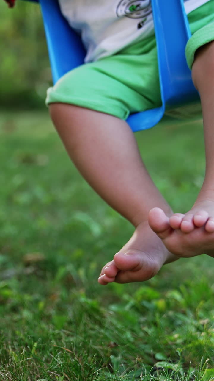 Little feet of a baby sitting on a swing. Barefoot child having fun outdoors. Close up. Blurred backdrop. Vertical video