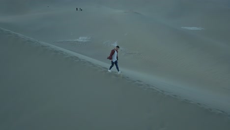 4k aerial follow shot of a boy walking alone in a barren desert on a large sand dune during a windy and dry day