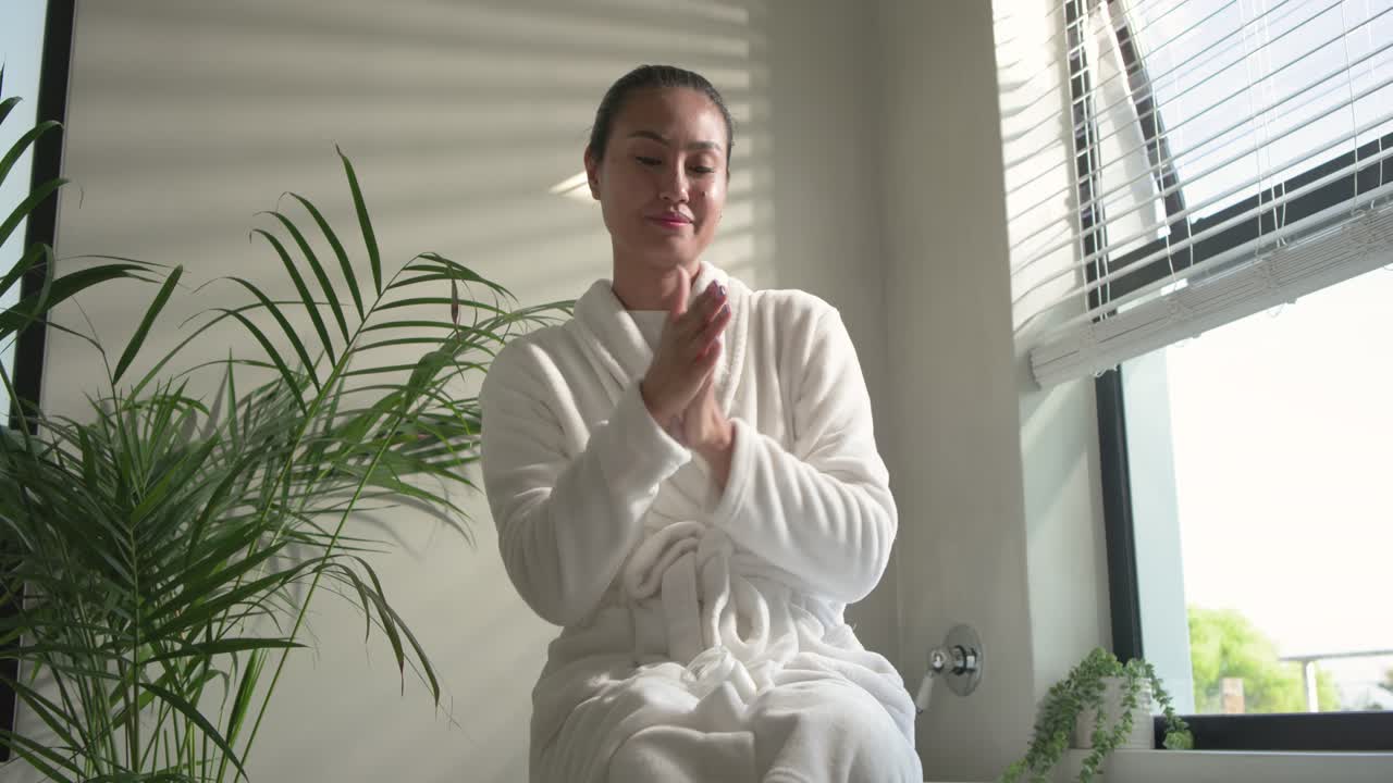 Mujer asiática presionando las manos contra el pecho y frotando una loción para el cuidado de la piel en el borde de la bañera, con la mirada fija