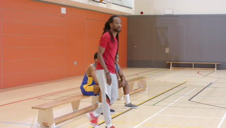 multiracial male basketball players in gym and with male coach observing game at bench