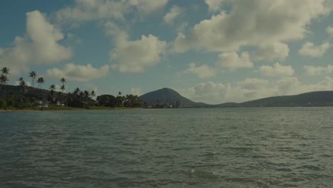 time lapse with the blue water of the pacific in the foreground and a volcanic island mountain and palm tree lined peninsula grace the horizon while white puffy clouds glide across the blue skies