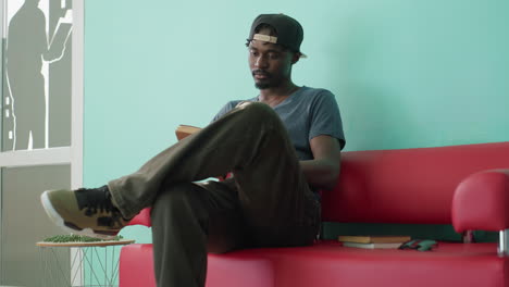 young student in casual clothing and cap stands near red couch, drops one book onto seat while opening another in hand, looking down with focus in bright library room with shelves and turquoise wall