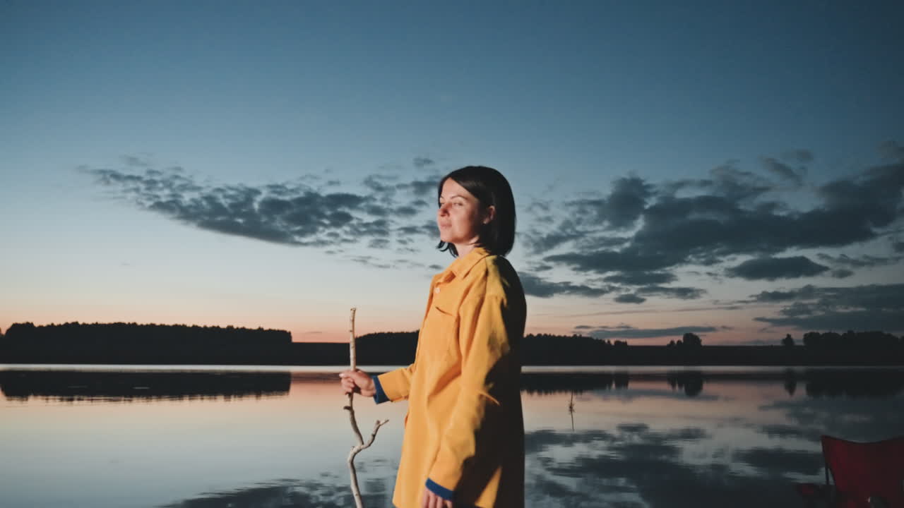 Kvinde ved søen under solnedgangscamping. En silhuet af en gul jakke ses på bredden vendt mod det rolige vand. En rød stol og fiskestang er i nærheden. Fredelig tusmørkehimmel med skyrefleksion og blid brise, en eftertænksom stemning.