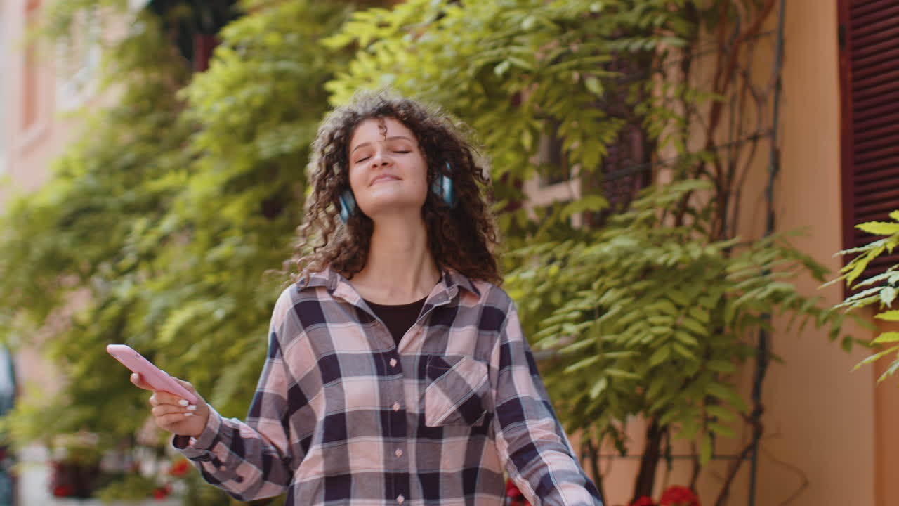 Happy young woman in wireless headphones choosing listening music dancing outdoors city street