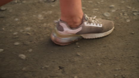 close-up of individual's legs walking along dirt road in rural environment, wearing casual sneakers, dusty gravel path visible, focus on movement