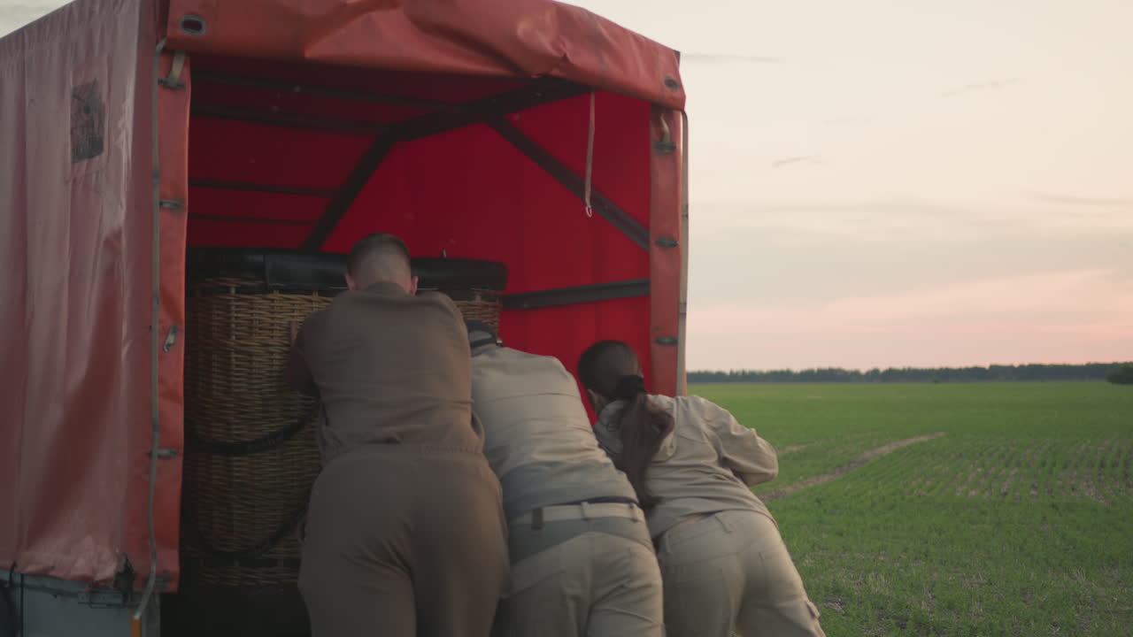 three people pushing wicker hot air balloon basket into truck trailer at dusk on open field, hands gripping basket rim and coordinating effort under soft sky, emphasizing teamwork