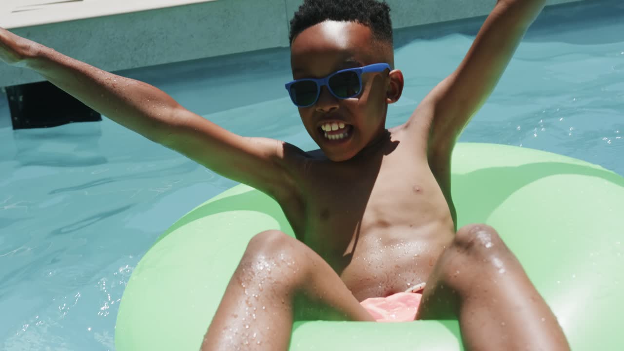 African boy kicking ring tilting, bracing backyard pool wall to steady in blue shades pink trunks