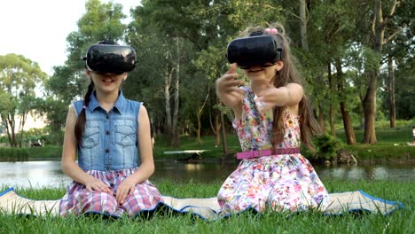 niñas felices se sientan en el parque en la hierba junto al río en gafas de realidad virtual y juegan juegos virtuales. ellos agitan sus manos y se ríen.