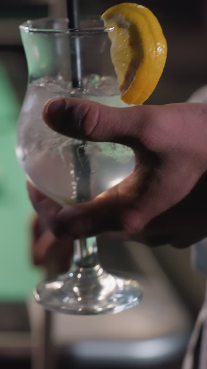 Primo piano della mano di una persona che tiene un bicchiere di bevanda al limone con cannuccia nera. Cubetti di ghiaccio e una fetta di limone aggiungono un dettaglio rinfrescante. Un tavolo da biliardo sfocato con bilie colorate crea un'atmosfera dinamica e rilassante.