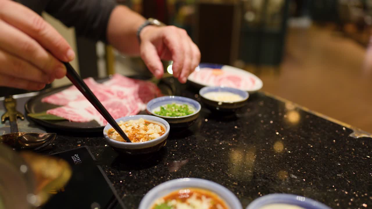 Hand combines garlic, scallions, and sauce for hotpot, under warm lighting at restaurant table