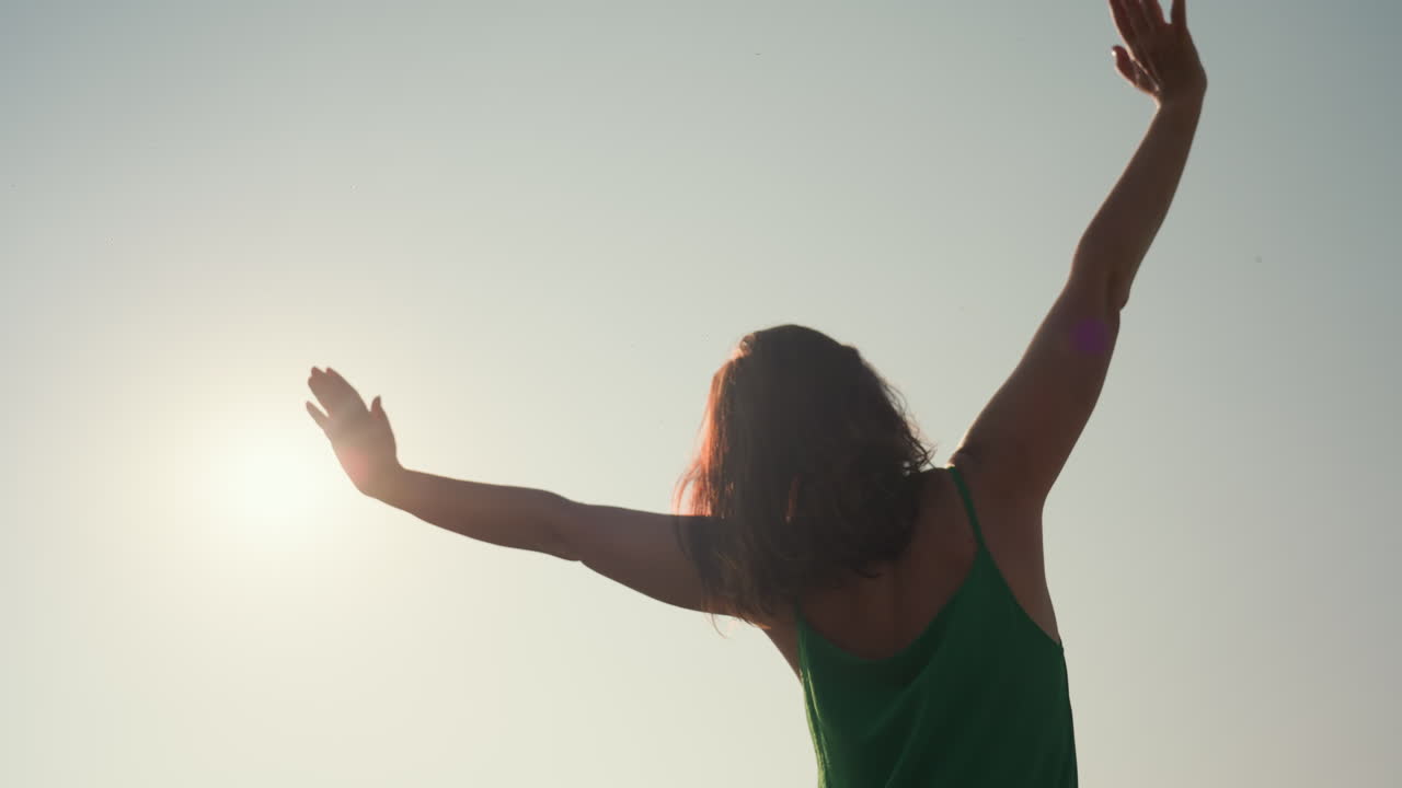 rear view of woman in green dress lifting hand to scratch hair while facing sun, sunlight casting warm glow around her silhouette under clear sky in peaceful outdoor scene during golden hour