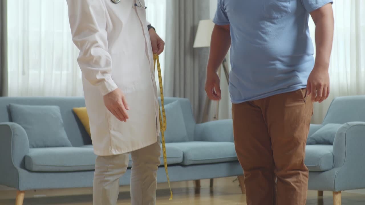 아시안 남성 의사가 클리닉에서 한 남성 환자를 가지고 있습니다. 환자는 무게를 측정하는 기계, 의사는 테이프를 사용하여 환자의 허리를 측정합니다.