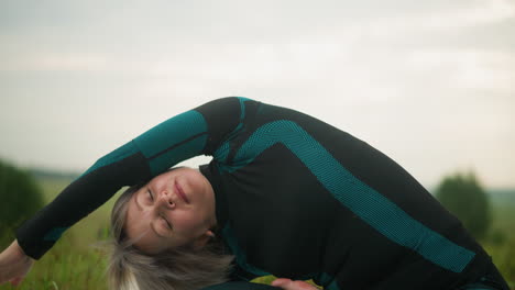 woman with grey hair lying on one side practicing side bend yoga pose with arm extended in a grassy field under a cloudy sky, background blurred