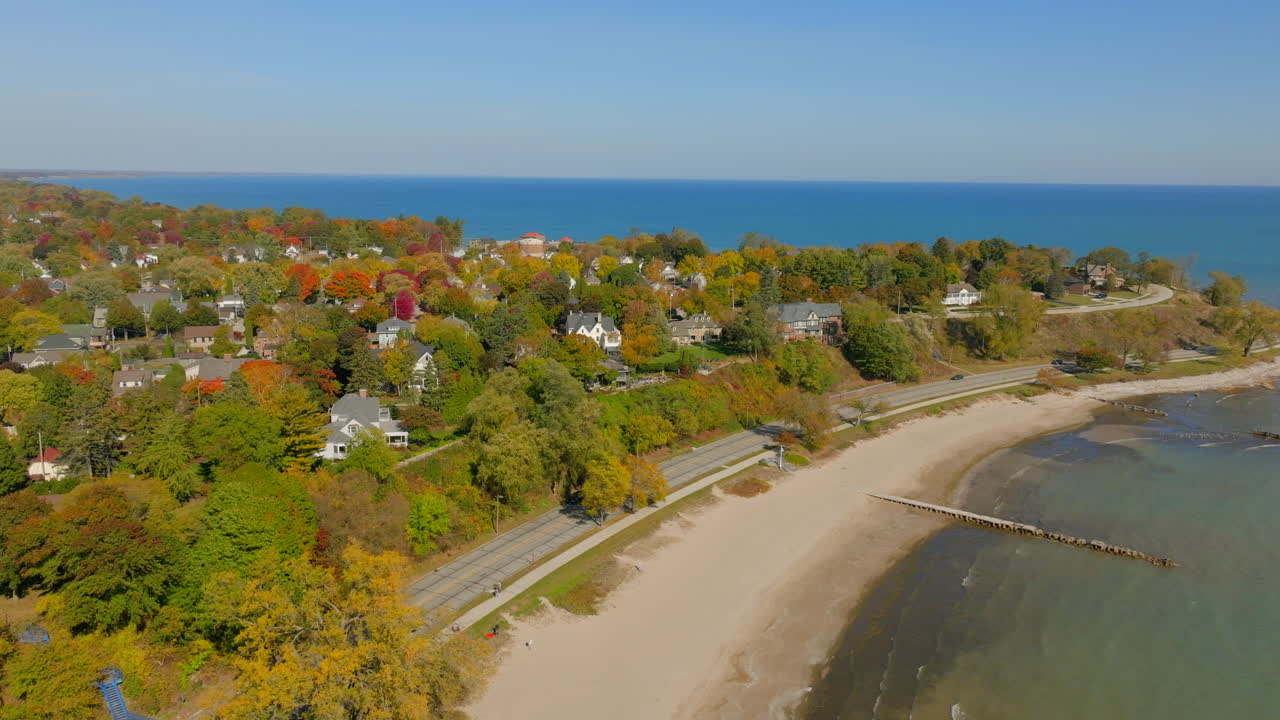 Drone aerial over Lake Michigan toward the Sheboygan, Wisconsin shoreline and road with charming neighborhood homes and colorful autumn trees beneath a clear blue sky