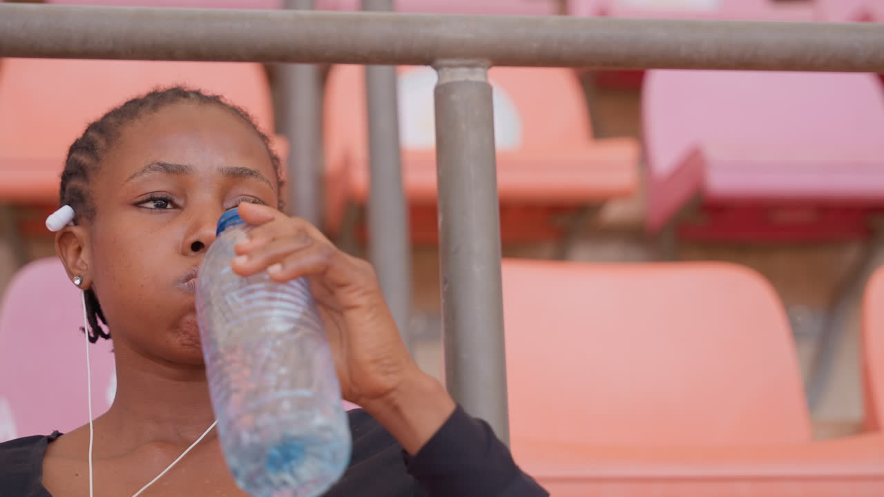 Spectator enjoys hydration during game moment. Closeup of fan drinking water with thoughtful expression. Individual with braided hair drinks water amid stadium seats with orange background