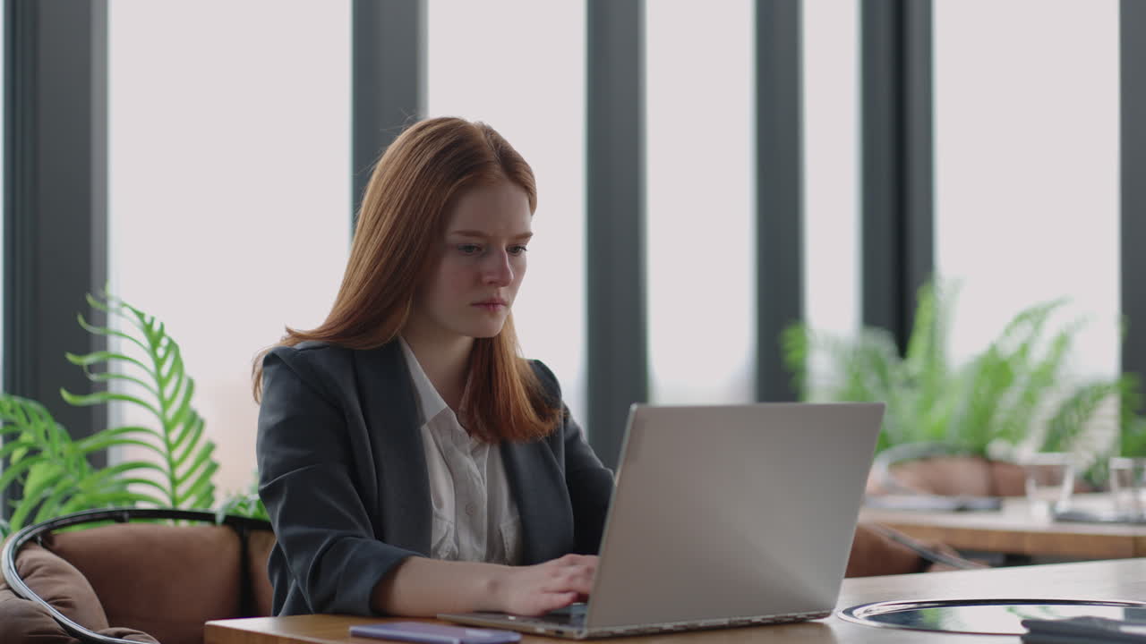 A young red-haired woman is working on a laptop. Remote work. Home office. Office work. A woman in a suit in the office is typing on a laptop