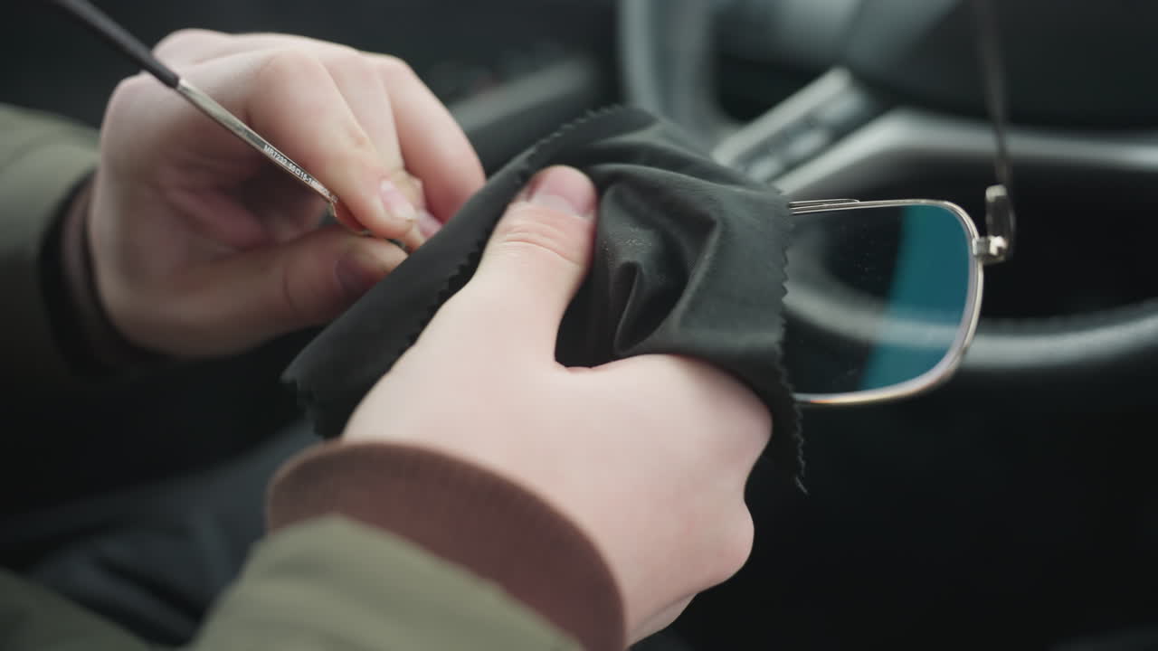 Close up of driver cleaning eyeglass lens with black microfiber cloth inside car cabin showing hand wiping sensitive optics near steering wheel under natural light capturing lens maintenance motion