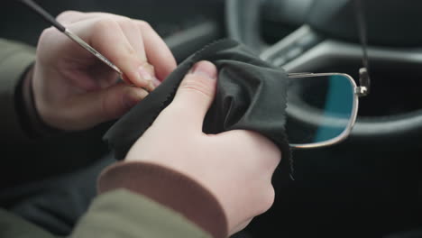 close up of driver cleaning eyeglass lens with black microfiber cloth inside car cabin showing hand wiping sensitive optics near steering wheel under natural light capturing lens maintenance motion
