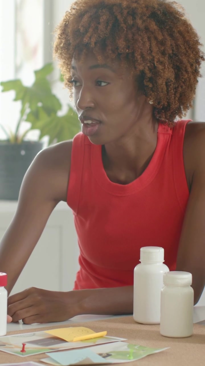 Vertical video: Spotting papers, woman in red top reviewing supplements near white bottles at home