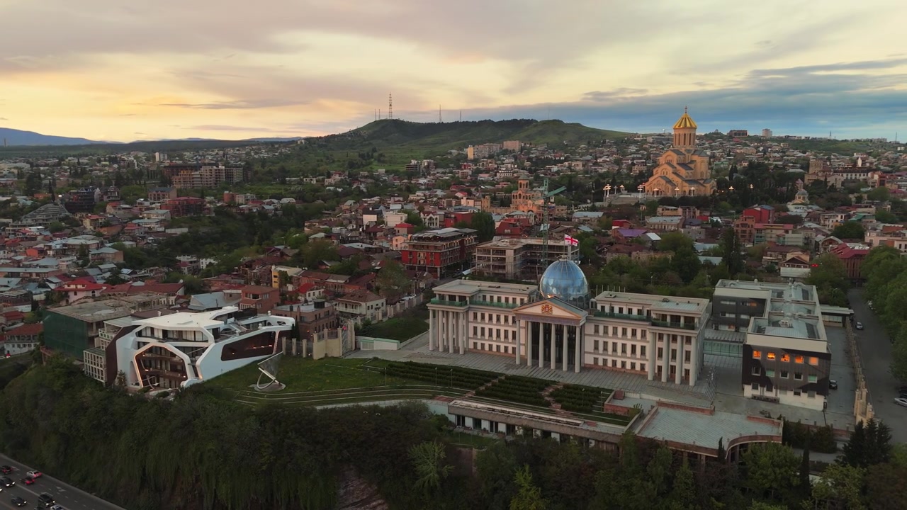 Letecký pohled na Tbilisi při západu slunce s prezidentským palácem s jeho skleněnou kupolí a katedrálou Nejsvětější Trojice (Sameba). Panorama gruzínské metropole s parkem Mtatsminda v pozadí.