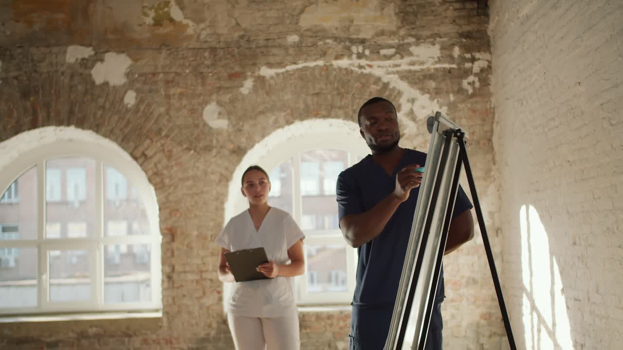 A Black male doctor in a blue uniform writes explanations on a canvas and addresses the audience, who listen and take notes as he leads a training session with his assistant, a female nurse in a white uniform, standing next to him in a spacious room with white brick walls