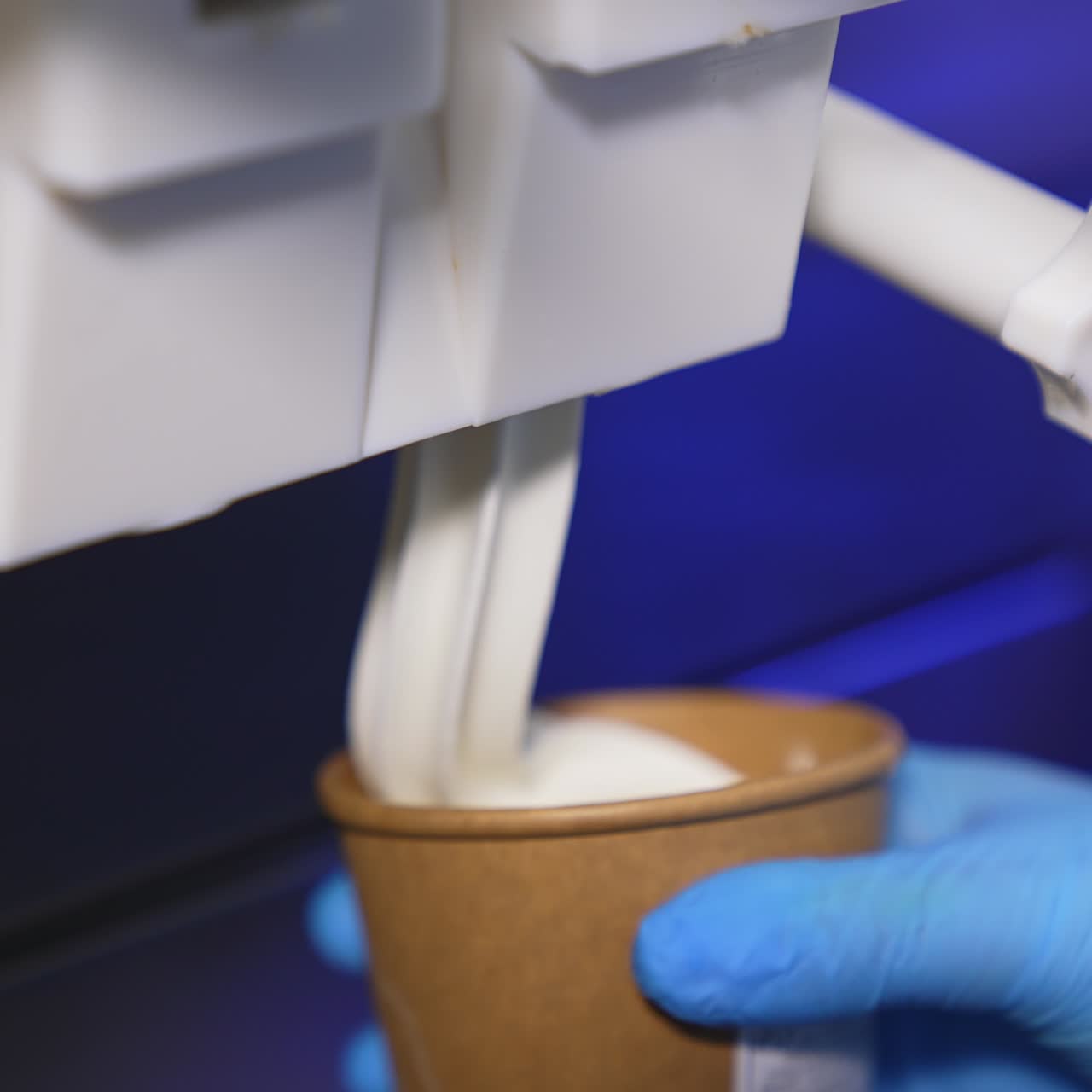 Hand in blue gloves puts a paper cup under a food automat. Soft creamy ice-cream is filling the cup close up