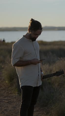 Man with Guitar and Smartphone Outdoors by the Water