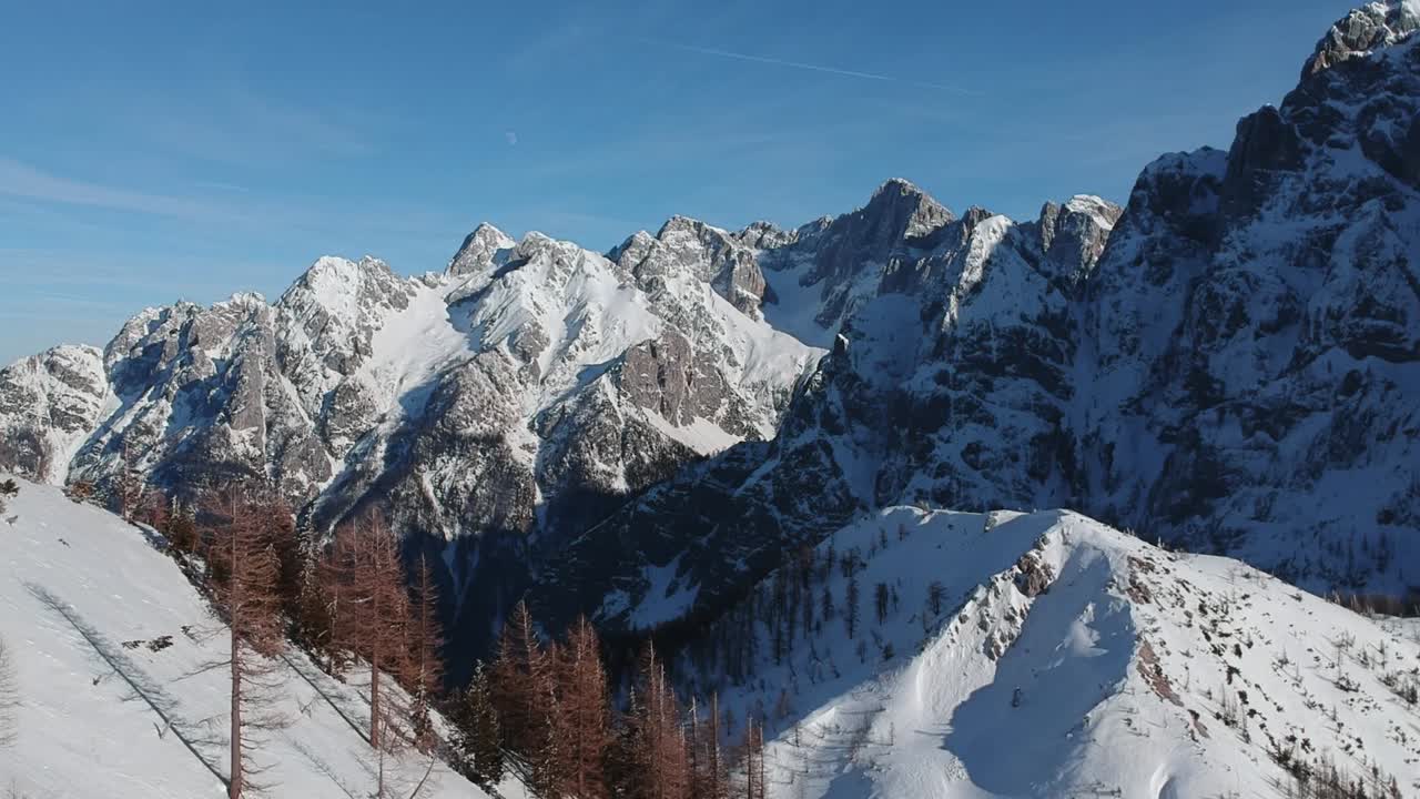 A snow covered rocky mountain range with pine trees beneath in Slovenia, mountain pass Vršič - aerial reveal shot