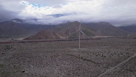 Slow-aerial-dolly-shot-of-a-wind-turbine-in-the-Palm-Springs-desert-on-a-cloudy-day-with-mountains-in-the-background-and-passing-cargo-train