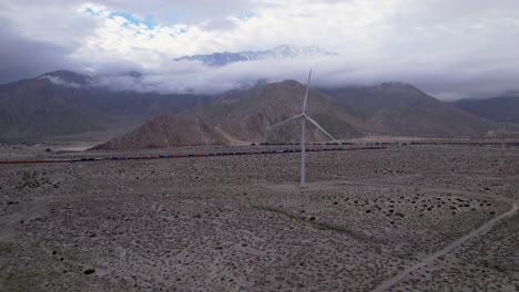 Toma-Aérea-Lenta-De-Una-Turbina-Eólica-En-El-Desierto-De-Palm-Springs-En-Un-Día-Nublado-Con-Montañas-En-El-Fondo-Y-Pasando-Un-Tren-De-Carga