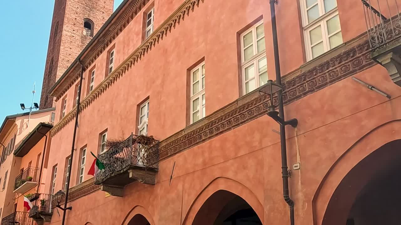 Un'inquadratura dal basso di una facciata in terracotta con finestre ad arco, balconi e bandiere italiane.