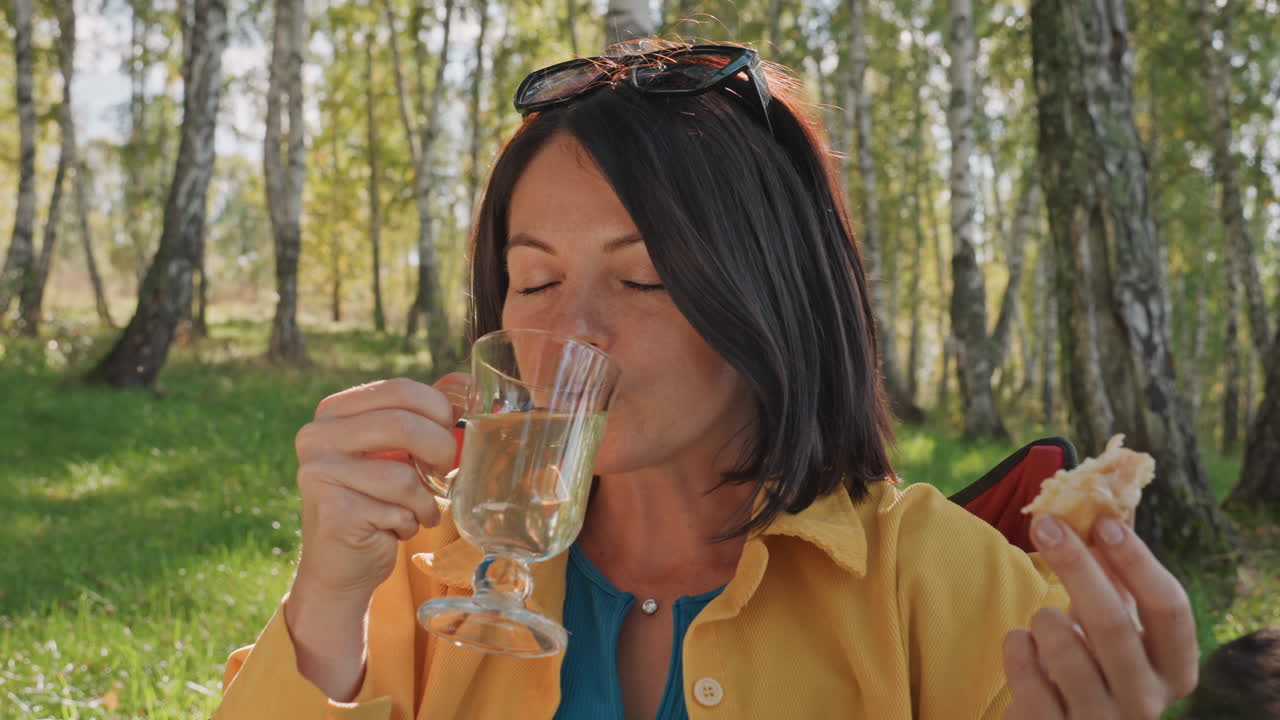 Donna rilassata che assapora un pasto sotto rami frondosi. Donna serena fa un picnic all'ombra degli alberi di betulla. Una donna sorride con calma mentre si gode il pasto all'ombra di alti rami di betulla.