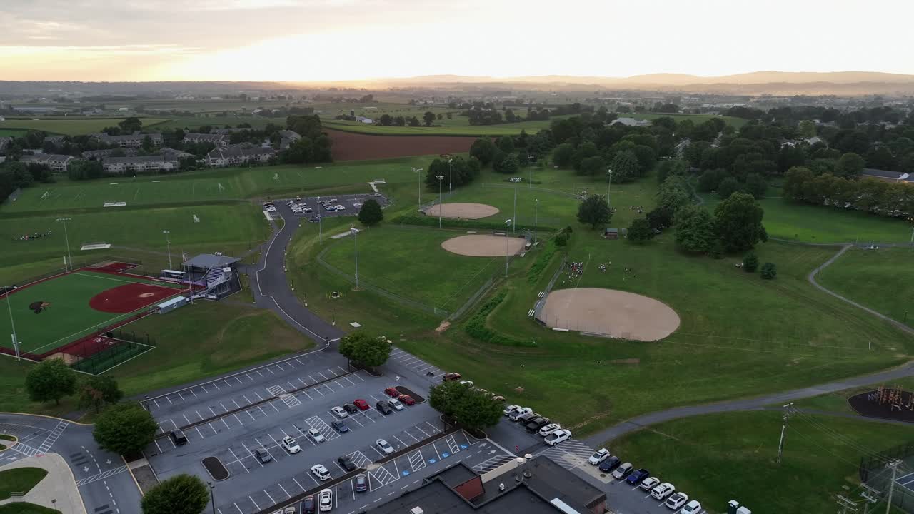 Aerial flyover American high school building with several baseball fields in background. Parking cars on lot. Foggy sunrise in the morning. Ephrata, Pennsylvania. Wide shot