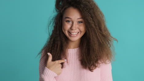 caucasian curly haired woman pointing at herself in front of the camera.