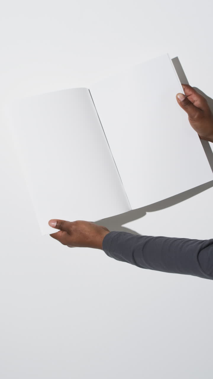 Vertical video of hands of african american man with book with white blank pages on white background