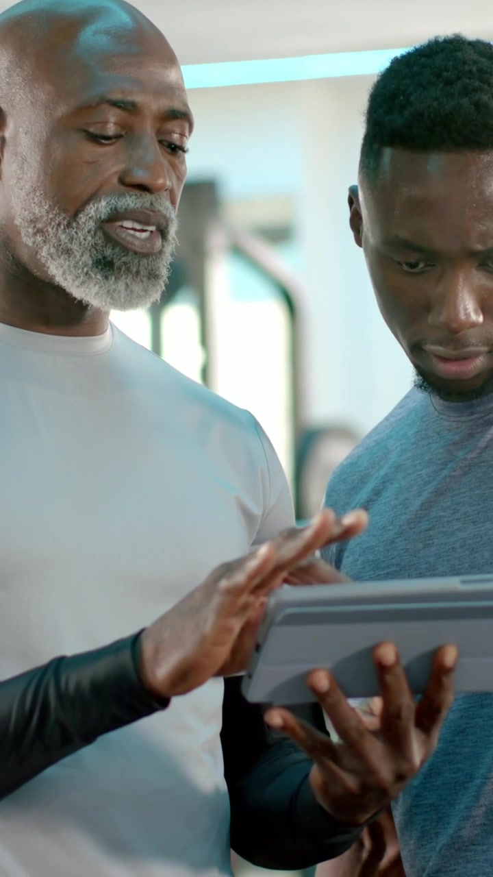 Vertical video: Tapping coach guiding sweaty trainee in gray tee at gym, pointing at tablet data