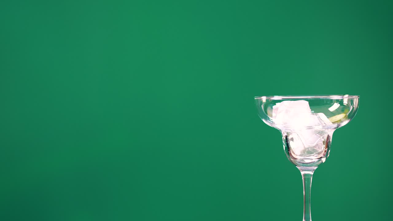 A cocktail glass with ice and lime slices against a green screen background. Hand adds lime slices with tongs