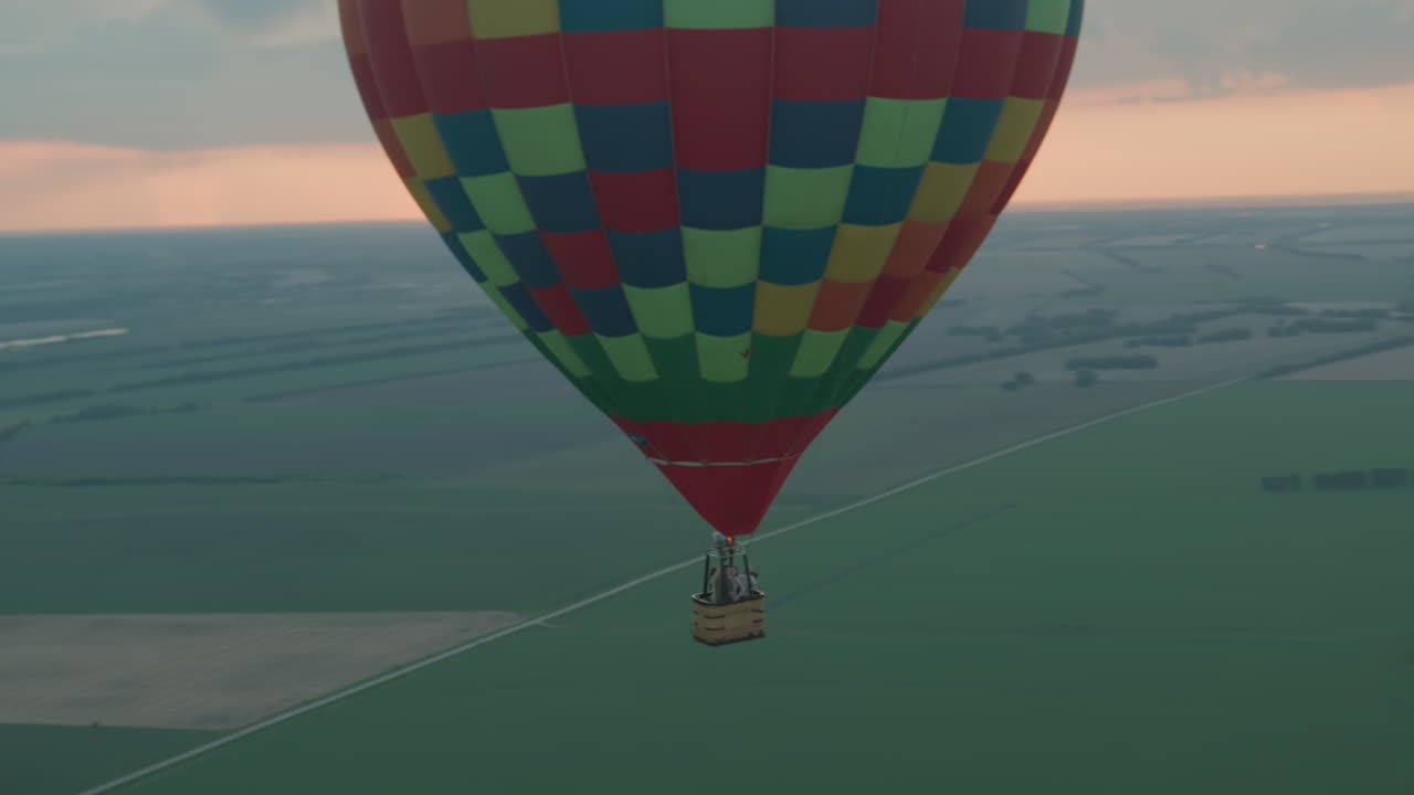 Colorful checkered hot air balloon carrying passengers drifting over vast farmland at sunrise, distant serene flight capturing panoramic aerial landscape, sky glowing pink under early morning clouds