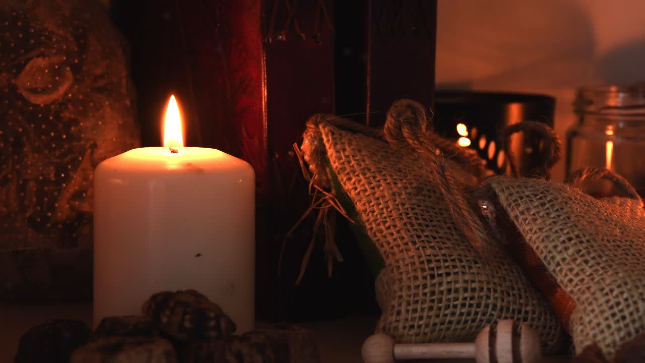 Avslappende detaljbilde i bakgrunnen av en urtetebutikk, med stearinlys med flakkende flammer, urter, bøker, små poser, krukker og litt støv som virvler rundt.
