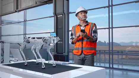 full body of asian male engineer with safety helmet using smartphone and looking around while standing in high tech factory, analyzing robotic machine concept