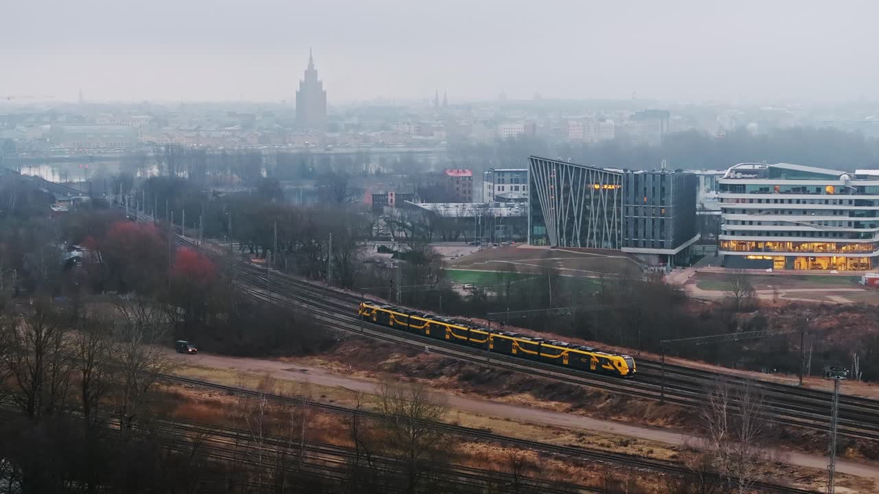 Yellow train travels through cold foggy cityscape in Latvia on February morning