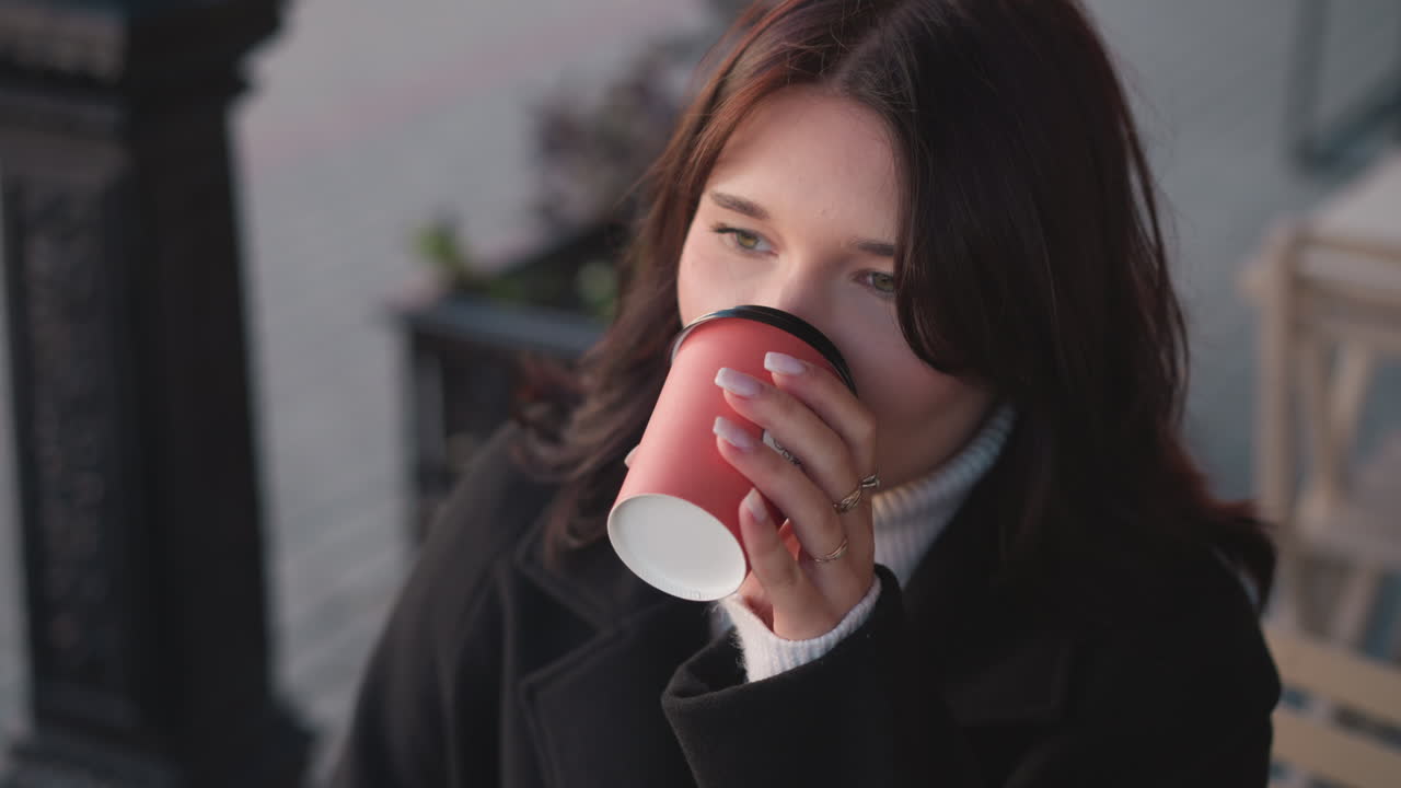 젊은 여자가 컵에서 커피를 마시며 웃으며 야외에서 시간을 즐기며 컵을 떨어뜨리고, 흐릿한 배경에는 지나가는 사람, 꽃, 장식 기둥이 있습니다.