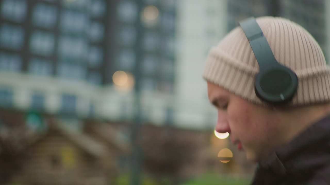 young white man dribbling basketball outdoors urban court near highrise buildings at dusk, wearing beanie and padded jacket with overear headphones, focused profile with motion blur, performing
