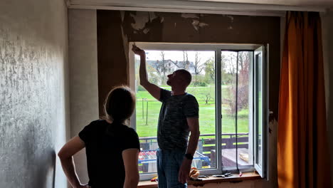 man applies adhesive on wall with brush while woman observes by window