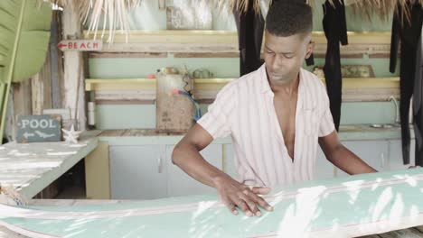 African-american-man-preparing-surfboard-on-the-counter-of-surf-rental-beach-shack,-slow-motion